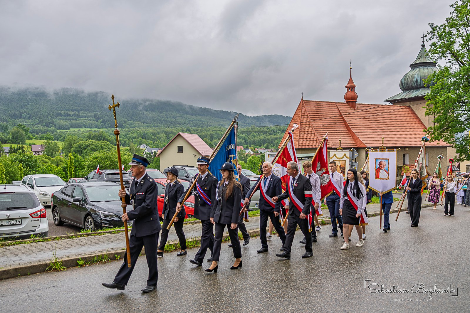 Procesja Bożego Ciała z chorągwiami na drodze w Bieńkówce, w tle kościół.