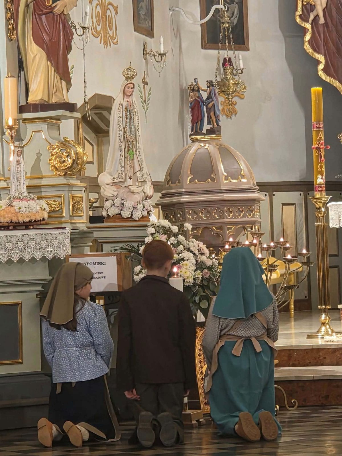 Dzieci w strojach pastuszków z Fatimy modlą się przed figurą Matki Bożej Fatimskiej.